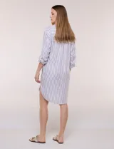 Vestido camisero corto con estampado a rayas verticales en color marrón y blanco, de cuello camisero con escote en V y botones frontales parciales. Cuenta con bolsillos laterales y en el pecho, mangas largas con presillas para ajustar y ruedo curvo asimétrico.