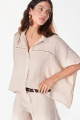 Camisa de lino color beige, de corte holgado y diseño corto, con cuello clásico, cierre frontal de botones y detalle de encajeado horizontal en el pecho y las mangas.