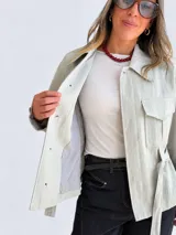 Campera blanca de algodón con cuello amplio, bolsillos delanteros con solapa, mangas largas y cinturón ajustable con hebilla redonda oscura. Cuenta con botones negros a contratono.