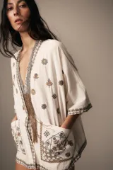 Blusa de escote en pico y manga corta, confeccionada en tejido color crudo con detalles de bordados florales y geométricos a contraste. Presenta cierre frontal con botones y bolsillos de plastrón en el delantero.