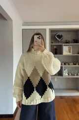 Buzo de tejido de punto grueso con cuello alto, mangas largas abullonadas y diseño de rombos en colores crema, beige y negro.