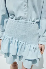 Pollera mini confeccionada en denim de algodón celeste, con cuerpo ajustado en punto smock y doble volante al bies en el bajo.