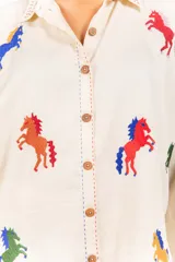 Camisa de corte clásico y calce amplio, color marfil, con cuello camisero, mangas largas y cierre frontal con botones. Presenta bordados coloridos de caballos en contraste distribuidos en la prenda.