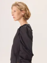 Camisa negra con mangas largas abullonadas y volados en los hombros y a lo largo de las mangas. El cuerpo de la prenda parece ser de un tejido más ligero y la espalda de algodón de remera.