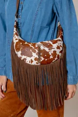 Bandolera de diseño medialuna confeccionada en cuero con pelo vacuno estampado tipo animal print manchado. Presenta un detalle de trenzado central, placa metálica con logo de la marca y flecos largos de gamuza marrón en toda la parte inferior. Incluye correa ajustable de cuero.