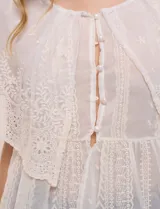 Blusa de estilo romántico color beige confeccionada en algodón, con escote amplio, importante volante capa en broderie calado, cartera frontal con botones tipo perla, bordados verticales calados, cintura marcada con bajo peplum y ruedo recto festonado.