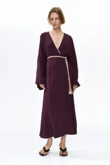 Vestido largo color bordó en tejido de viscosa, modelo oversized con escote en V cruzado y ribetes en contraste. Presenta mangas largas amplias, costura fruncida en la cintura y un cinturón trenzado con borlas y cuentas en los extremos.