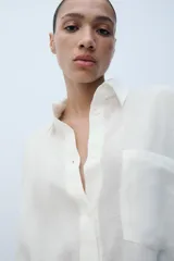 Camisa oversize blanca de cuello solapa y manga larga con puños anchos. Cuenta con cierre frontal de botones y un bolsillo aplicado en el frente.
