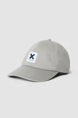 Gorra de béisbol color gris claro, con visera curva y parche frontal cuadrado con una 'X' azul marino y texto 'blue banana' en la parte inferior.