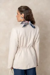 Chaquetón color beige de corte recto y largo, con cierre frontal mediante botones y lazo ajustable en la cintura. Presenta cuello en V, bolsillos laterales con solapa y mangas largas.