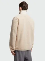Buzo de polar tipo teddy en color beige, con cierre frontal completo, cuello alto y bolsillo en el pecho con solapa y logo de la marca. Presenta detalles en tela técnica al tono en los bordes de los bolsillos, cierre y cuello.