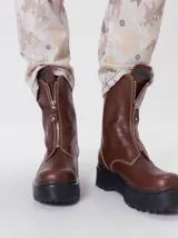 Botas de cuero color marrón de caña alta, con plataforma gruesa de goma y cierre metálico frontal con tirador. Presentan costuras a contratono y detalle de etiqueta de la marca en la lengüeta.