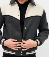 Campera de estilo varsity confeccionada en polyvelour, con diseño bicolor en gris oscuro y crema. Presenta cuello tipo polo, cierre frontal con botones a presión, mangas largas y puños y cintura con terminación en ribana a rayas.