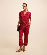 Blusa de crepé color rojo con estampado geométrico de rombos en negro y blanco. Tiene cuello redondo con abertura en forma de gota y detalle metálico, mangas cortas y corte recto.