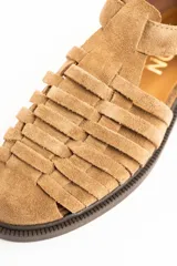 Sandalias cangrejeras de gamuza color visón, con diseño de tiras entrelazadas, tira ajustable con hebilla metálica en el tobillo y suela plana de goma marrón.