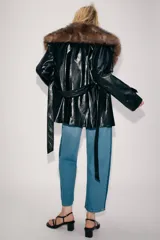 Trench corto Zara de efecto piel color negro, con cuello de solapa y aplicación de pelo sintético a contraste en color marrón. Cuenta con manga larga con trabillas en los puños, bolsillos delanteros y cinturón ajustable del mismo tejido.