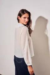 Blusa blanca de algodón peinado con mangas largas, cuello babero bordado y pliegues frontales.