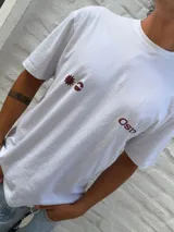 Remera blanca de corte oversize con cuello redondo y manga corta. Presenta un estampado frontal en color marrón rojizo con un diseño abstracto que incluye un sol estilizado y la palabra "Osten".