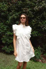 Vestido corto de lino color blanco, con cuello alto fruncido, volados marcados en los hombros y caída recta. Presenta botones en el frente.