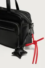 Bolso bandolera negro de acabado liso, con compartimento principal con cierre de cremallera. Cuenta con dos bolsillos frontales, uno con solapa y detalle metálico y otro con cremallera. Incluye dos asas de mano y un asa de hombro ajustable y extraíble, además de detalles de charms y mosquetón en un lateral.