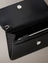 Billetera tipo sobre de Calvin Klein, color negro, confeccionada en piel sintética. Presenta el logo 'ck' grabado en el frente, cierre con solapa y correa desmontable, permitiendo su uso como cartera pequeña o clutch.