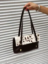 Cartera de hombro combinada en cuero liso marrón oscuro y cuero con pelo vacuno estampado tipo animal print. Presenta solapa frontal con dos correas decorativas con hebillas metálicas y detalles de herrajes en bronce.