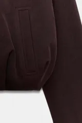 Chaqueta estilo bomber de color marrón oscuro, con cuello redondo, manga larga y cierre frontal con cremallera. Presenta bolsillos delanteros de vivo y un diseño con bajo fruncido.