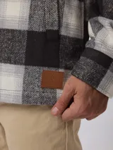 Sobrecamisa de estilo leñador con estampado de cuadros en tonos gris, negro y blanco. Presenta cierre frontal con cremallera y solapa con botones a presión, cuello clásico y bolsillos laterales.
