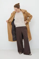 Trench coat color beige con cuello de solapa, cierre frontal abotonado, bolsillos laterales y mangas largas con cintas ajustables. Incluye capucha desmontable y forro interior rayado. La tela exterior cuenta con protección repelente a la lluvia y manchas.