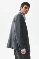 Blazer cruzado de color gris medio, con solapas de pico, ojal decorativo y cierre de doble botonadura. Presenta bolsillo superior insertado, bolsillos delanteros ribeteados con solapa y botones decorativos en los puños.