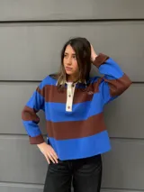 Buzo tipo rugby de algodón con diseño de rayas horizontales anchas en azul y marrón. Presenta cuello polo en color crema con cierre frontal de tres botones y mangas largas.