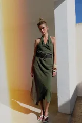 Vestido midi verde militar de cuello halter con escote en V cruzado y espalda descubierta. Cuenta con pliegues laterales, bajo asimétrico y un cinturón incorporado con hebilla redonda oscura.