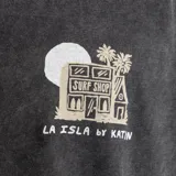 Remera negra de algodón con estampa en el pecho de una casa de surf, una palmera y el texto "La Isla by Katin".