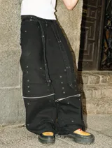 Pantalon baggy-flared de denim negro con efecto gastado, diseño desmontable con cierres horizontales en las piernas. Presenta múltiples parches de refuerzo, apliques metálicos, ojales decorativos y correas ajustables en los tobillos. Incluye un cinturón textil con tachas.