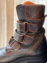 Botas altas de cuero marrón con suela track y tres tiras con hebillas metálicas en la caña.