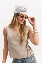 Chaleco beige con sutiles rayas verticales oscuras, escote en V cruzado y cierre lateral con lazo ajustable.