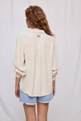 Camisa de manga larga color beige con cuello clásico y botonadura frontal. Presenta mangas con volumen sutil, puños abotonados y una silueta amplia de caída fluida.
