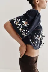 Top corto de color negro con escote en pico y mangas cortas anchas. Presenta bordados coloridos en estilo geométrico y floral, detalle de entredoses y cierre frontal mediante lazos con abalorios.