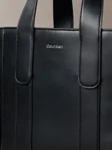 Cartera tote estructurada Calvin Klein de piel sintética lisa en color negro, con asas superiores de mano, compartimentos exteriores en los laterales y logo metálico grabado en la parte delantera.