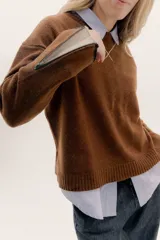 Buzo de lambswool color beige, con escote redondo y detalle en V. Presenta cierres metálicos en los puños que revelan un interior en contraste de color marrón y ribete rojo. Tiene tajos laterales para mayor comodidad y un fit amplio.