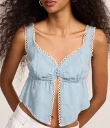 Top cropped de jeans celeste con escote corazón, breteles anchos y abertura frontal central abotonada. Los bordes del escote y la abertura están decorados con un ribete de encaje blanco tipo picot.