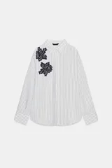 Camisa de cuello solapa y manga larga, con estampado de rayas verticales blancas y grises. Presenta dos aplicaciones de flores de encaje negro en la parte frontal. Cierre frontal con botones ocultos por solapa.