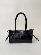 Cartera de cuero de tamaño medio con estructura rígida. Presenta una tira decorativa con tachas y hebillas metálicas en los extremos, además de lazos laterales. Cuenta con doble asa para llevar al hombro y cierre superior.