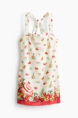 Vestido mini de color beige con un estampado de tomates, aceitunas y botellas de aceite, y un borde inferior rojo con ilustraciones de una sombrilla de playa y vegetales. Cuenta con escote cuadrado y tirantes anchos cruzados en la espalda.