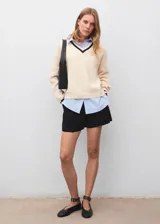 Jersey de punto grueso en color crema con cuello en V y detalles en contraste oscuro en el cuello y puños. Presenta un diseño recto de manga larga con una camisa de cuello camisero integrada en color celeste.