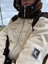 Campera de gabardina con diseño bicolor en tonos crudo y chocolate, con capucha forrada en polar. Presenta cierre frontal, mangas largas con puños elásticos y un parche distintivo en la manga.