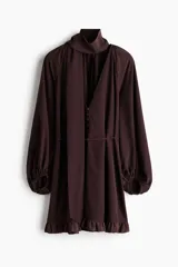 Vestido oversized corto de color marrón oscuro, con escote en V pronunciado y pañuelo haciendo juego que puede abrocharse en la nuca. Cuenta con botones forrados adelante, cinturón fino para atar, mangas globo largas de corte raglán con elástico en los puños y ribete de volados en el bajo.