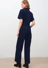 Mono largo de denim color azul oscuro, con cuello camisero, cierre frontal con cremallera y mangas cortas. Cuenta con bolsillos de parche delanteros y cinturón ajustable del mismo tejido.