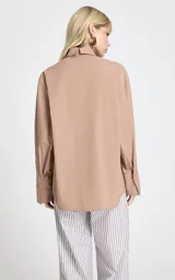 Camisa de corte recto y diseño oversize, con estampado de rayas verticales en tonos marrón y blanco. Presenta cuello clásico estructurado, mangas largas con puños anchos, hombros caídos y bajo asimétrico con abertura lateral. Cierre frontal mediante botones.