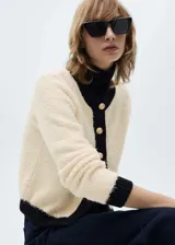 Chaqueta de punto de textura suave en color crema con detalles en contraste negro en cuello, puños, ruedo y tapeta de botones. Presenta un diseño recto, cuello redondo y cierre frontal con botones dorados.